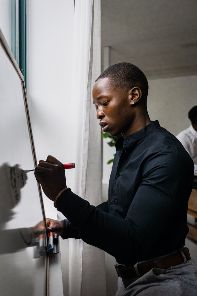 Black male in black shirt writing on a whiteboard indoors.